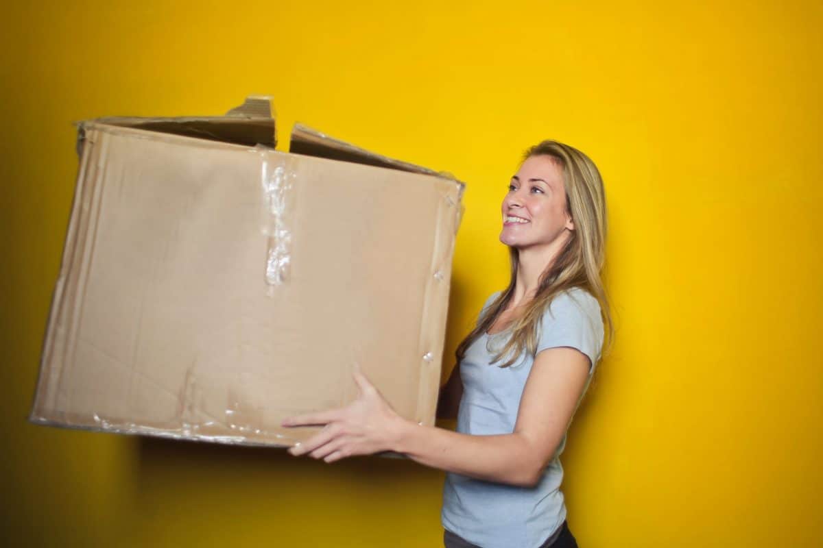 A woman in a gray shirt moves a large box in front of a yellow wall. 