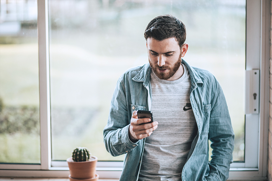 A man in his thirties is looking through his phone while standing in front of a window and a small potted succulent.