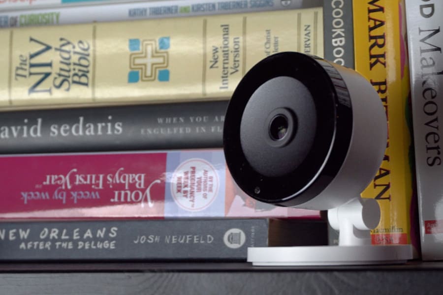 A white Alarm.com indoor camera is on a bookshelf in front of a stack of books.