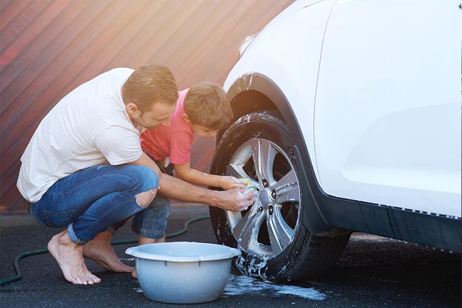 A man and his son are using a bowl of soapy water to wash a white car.