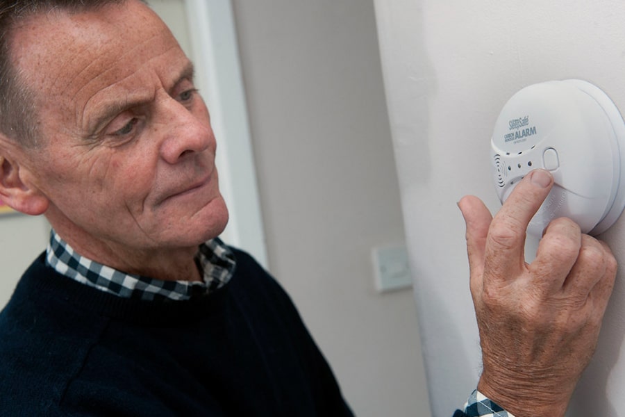 An older man wearing a dark sweater is adjusting a white carbon monoxide detector inside his home.