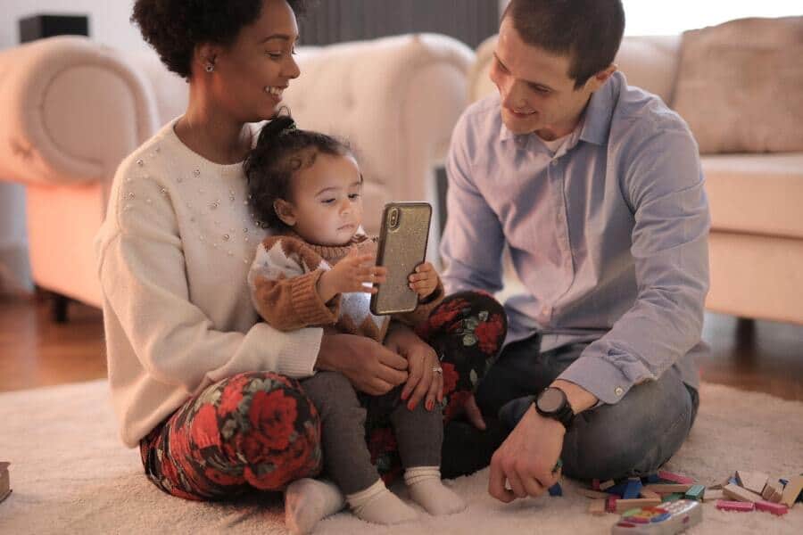 A toddler sitting on her mother’s lap holds her mother’s smartphone as the mother and father look on while sitting on a rug in their living room. 
