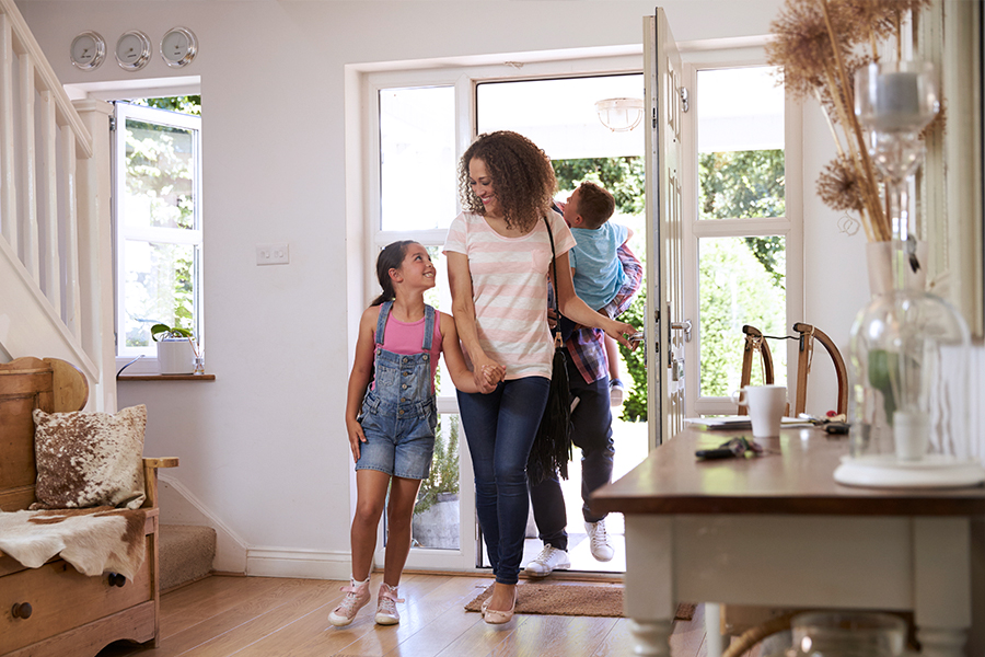 A mother and her young daughter hold hands as they enter their home through the front door while the father carries their young son behind them. 