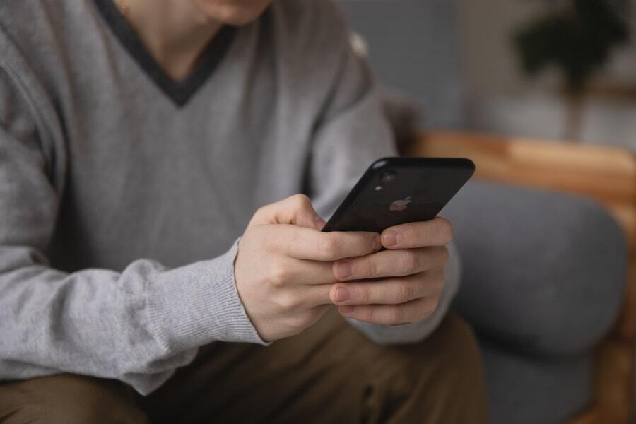 A man wearing a grey sweater is using his black iPhone while sitting on a grey sofa inside of his home.