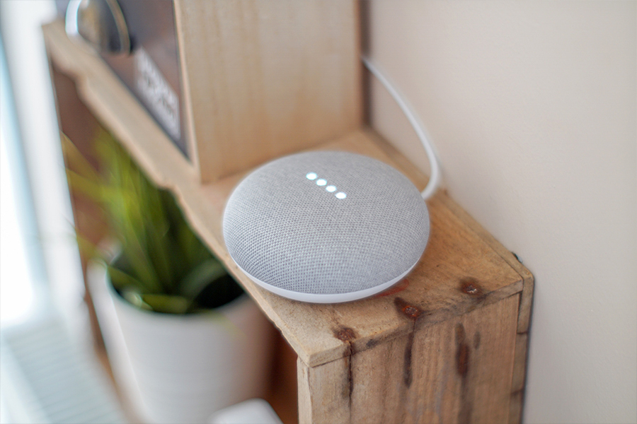 A grey and white Google Home Mini sits on top of a brown wooden bookshelf that is also holding a plant and other miscellaneous items.